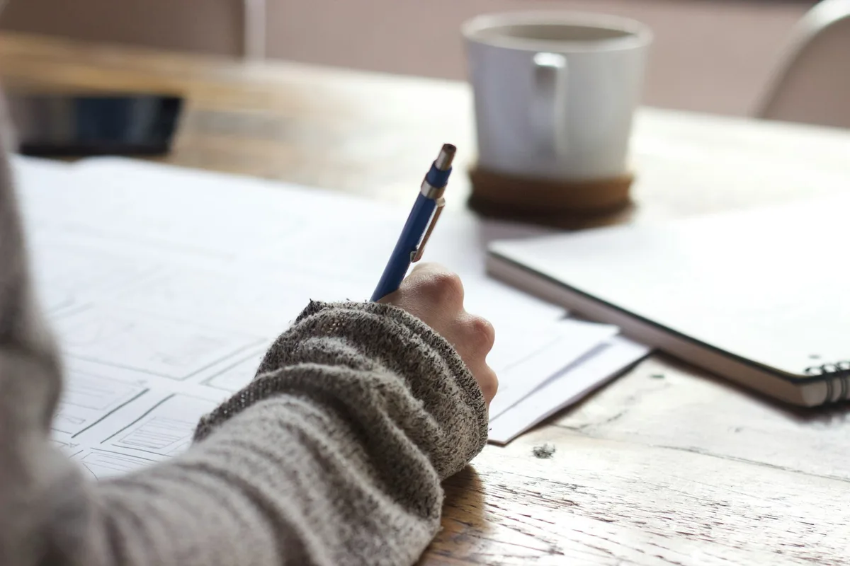 A person writing by hand at a wooden table near a mug.